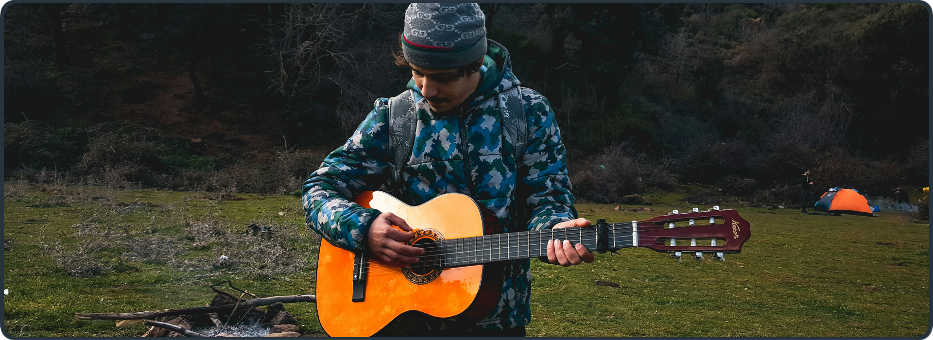 Junger Mann spielt Gitarre vor Zelt und Lagerfeuer – Symbol für verschiedene Kompetenzen im Studium